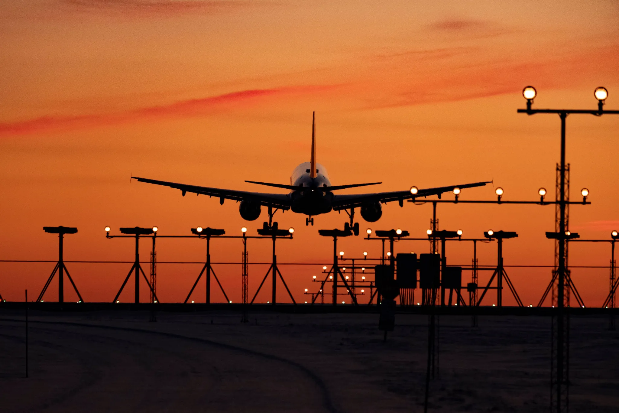 An airliner on final approach at sunset, silhouetted above a row of runway approach lights