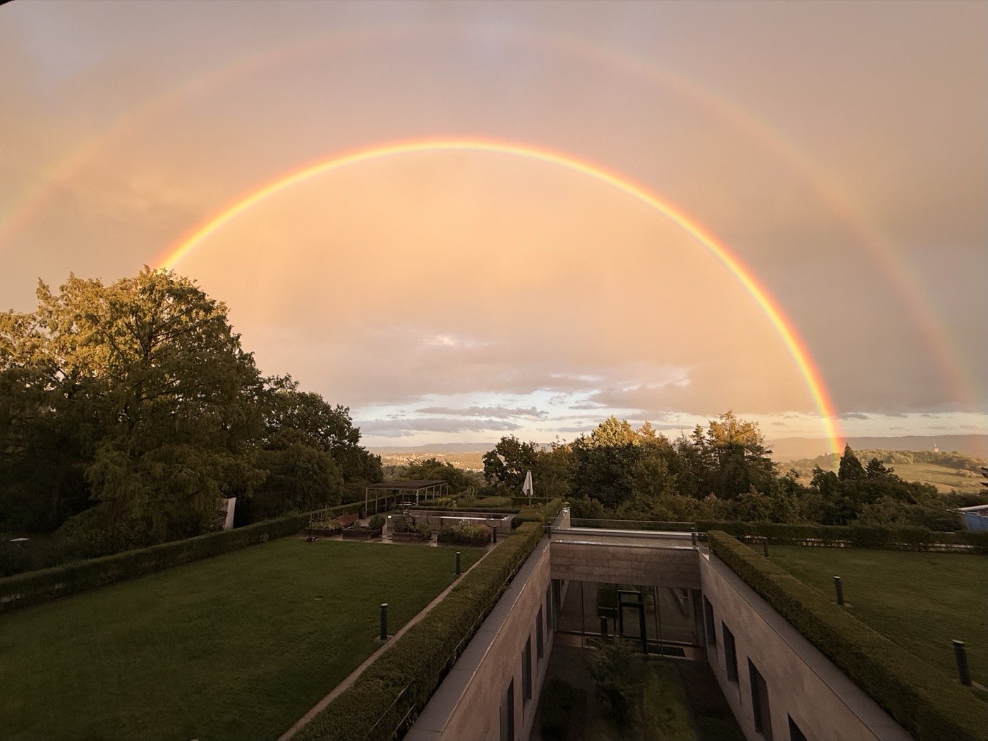A double rainbow arcing over a low modern building and the hills beyond, at sunset